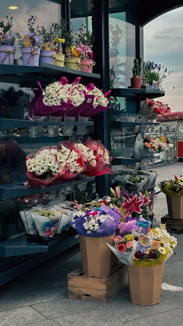A display of various potted flowering plants, including purple and white blooms, arranged on a black metal shelf inside a greenhouse or flower shop. The scene is framed by lush green ivy climbing up t