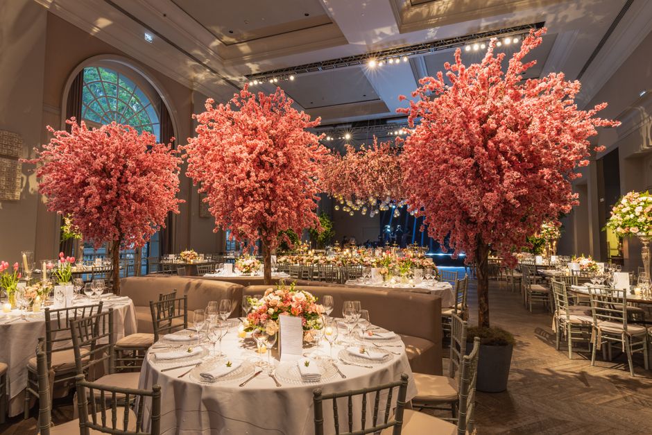 An elegant indoor banquet hall decorated with large, vibrant pink cherry blossom trees placed among round tables. The tables are draped with white tablecloths and set with fine glassware, cutlery, and