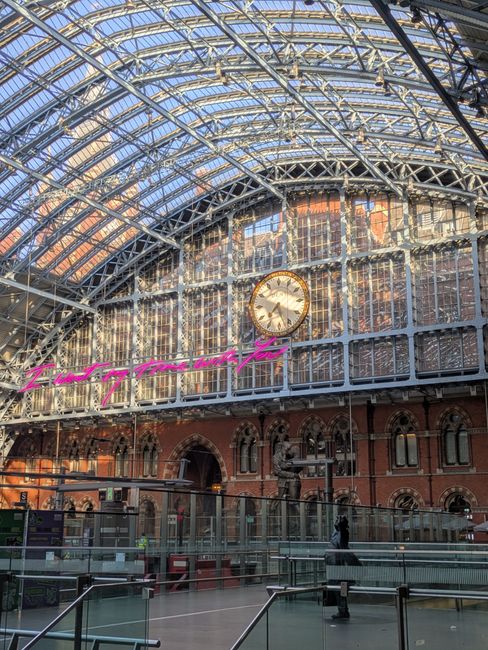 A large, arched glass ceiling at St Pancras station featuring intricate metal framework. Below the ceiling, a vintage-style clock with a white face and black numerals is mounted on a brick wall. Soft 
