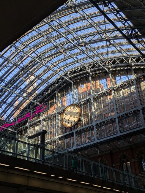 A large, arched glass ceiling at St Pancras station featuring intricate metal framework. Below the ceiling, a vintage-style clock with a white face and black numerals is mounted on a brick wall. Soft 