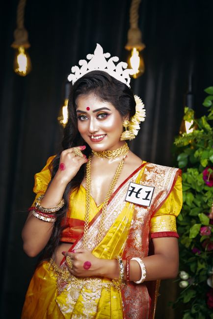 A young woman dressed in traditional Indian attire, wearing a vibrant yellow saree with intricate gold and orange embroidery, and accessorized with gold jewelry including a choker, earrings, and bangl