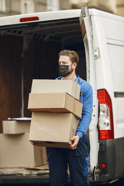 A young male delivery courier with neatly styled light brown hair, wearing a black face mask and a bright blue jacket, stands beside an open white delivery van holding three cardboard boxes stacked in
